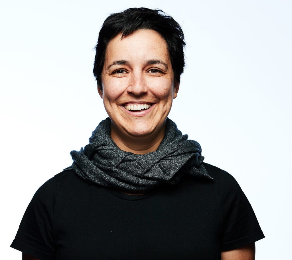 Headshot of Gabriela de Queiroz, a Latina with short black hair and brown eyes, a black shirt, and a gray neck scarf, smiling at the camera.