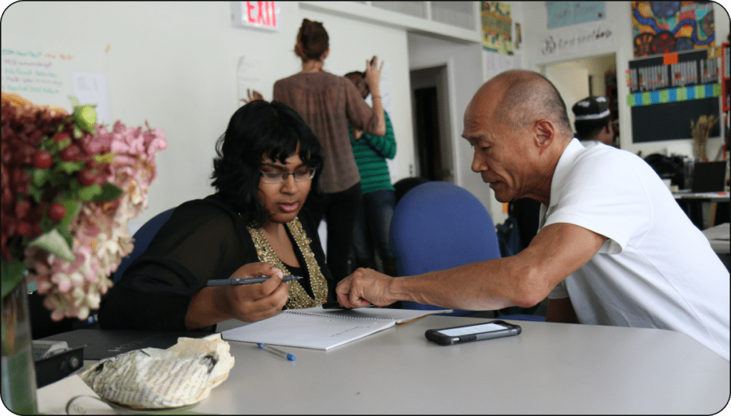 A middle-aged Asian man works with a younger Asian woman at a table. He's pointing to something in her notebook. They are working in a collaborative workspace with other people with underrepresented identities.