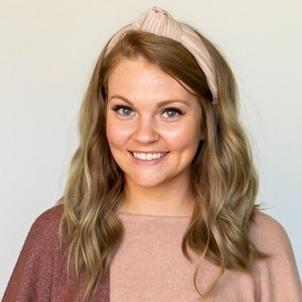 Headshot of Carly, a White woman with dirty blonde hair, a cream-colored fabric headband, and a color-blocked mauve and blush top smiling at the camera in front of a white wall.