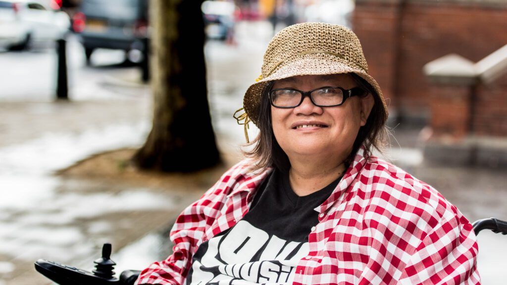 of Eleanor Lisney, an East Asian woman with short brown hair, glasses, a woven hat, a black t-shirt, and red and white plaid flannel