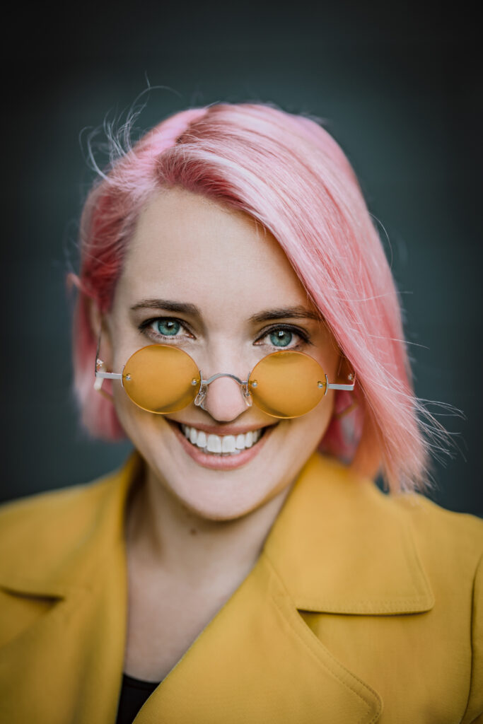 Headshot of Aubrey Blanche, a Latina with pink hair, blue eyes, glasses, and a yellow shirt.
