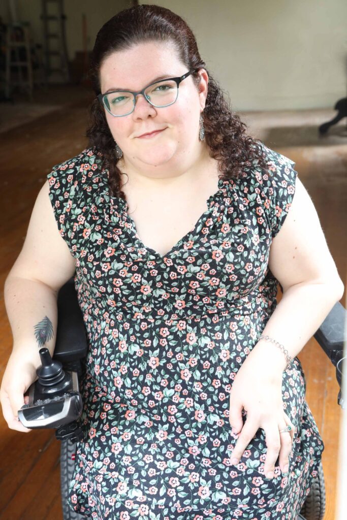 Headshot of Emily Ladau; a White woman who is wearing glasses and has curly brown hair pulled back halfway, with the rest framing her face. She is wearing a black dress dotted with red flowers and green leaves. She is sitting in a power wheelchair, facing the camera, and smiling. One of her hands is resting in her lap and the other hand is resting on the joystick of her wheelchair, turned so that a tattoo of a peacock feather is visible on her inner arm.