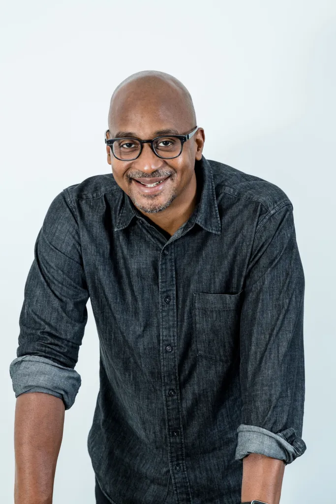 Professional photo of Wayne, a bald Black man with glasses, a goatee, and charcoal button-down leaning forward on a table in front of a white wall