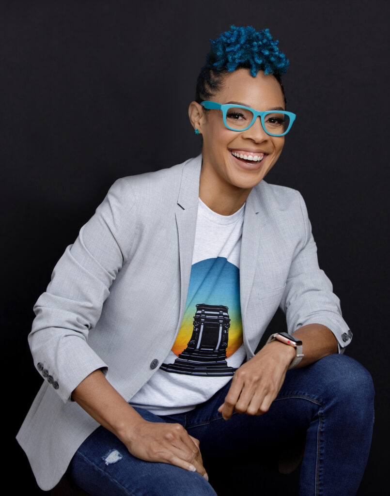 Headshot of Doc Jana, an African-American nonbinary person with short curly blue hair, matching blue glasses, a gray suit on top of a blue and white t-shirt, and an electric smile