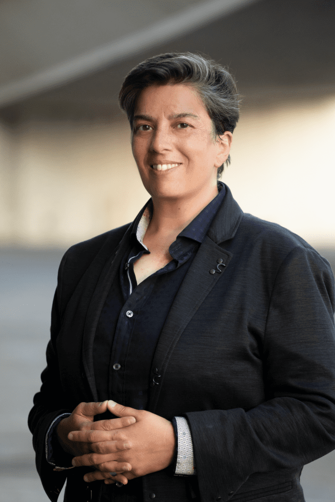 Headshot of Rajkumari Neogy, a French-Canadian/East Indian nonbinary person with short salt and pepper hair and a navy blue button-down with a black suit jacket, holding hands and smiling at the camera.