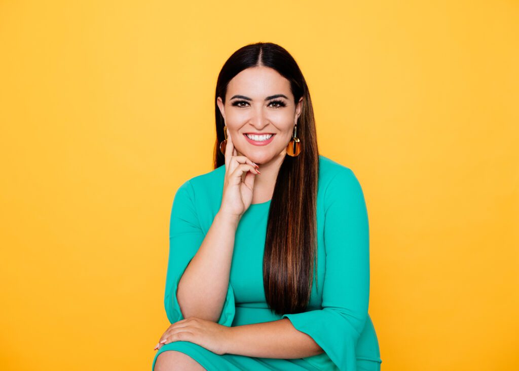 Headshot of Gabriela Chavez-Lopez, a Latina with long brown hair, chunky gold earrings, and a bright aqua dress, smiling to the camera in front of a bright yellow wall.