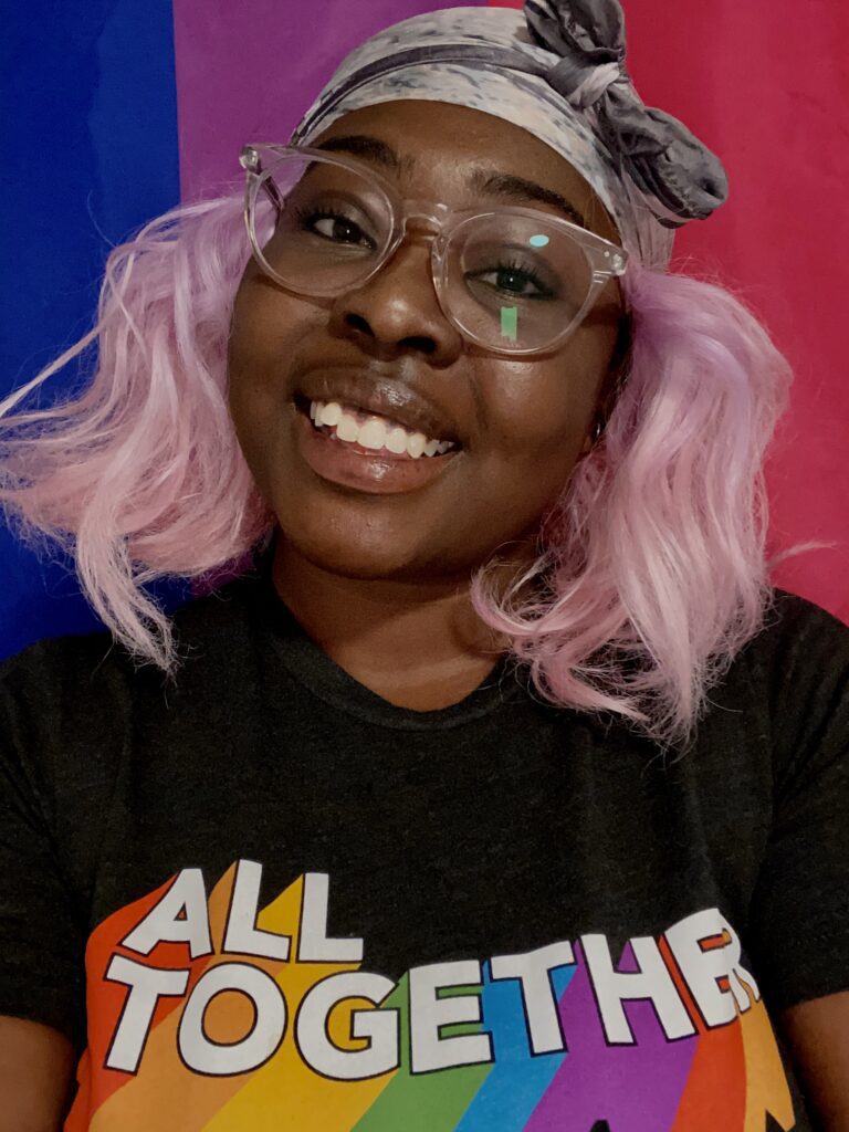 Headshot of Ashantè Fray, a Black bisexual woman with short pink hair, a ribbon head scarf, glasses, and a black shirt with the inscription "All together" in colors, smiling at the camera.