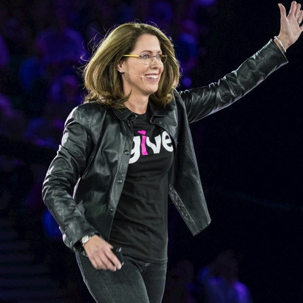 Photo of Kate Johnson, a White woman with brown hair, glasses, and a leather jacket, waving to the audience