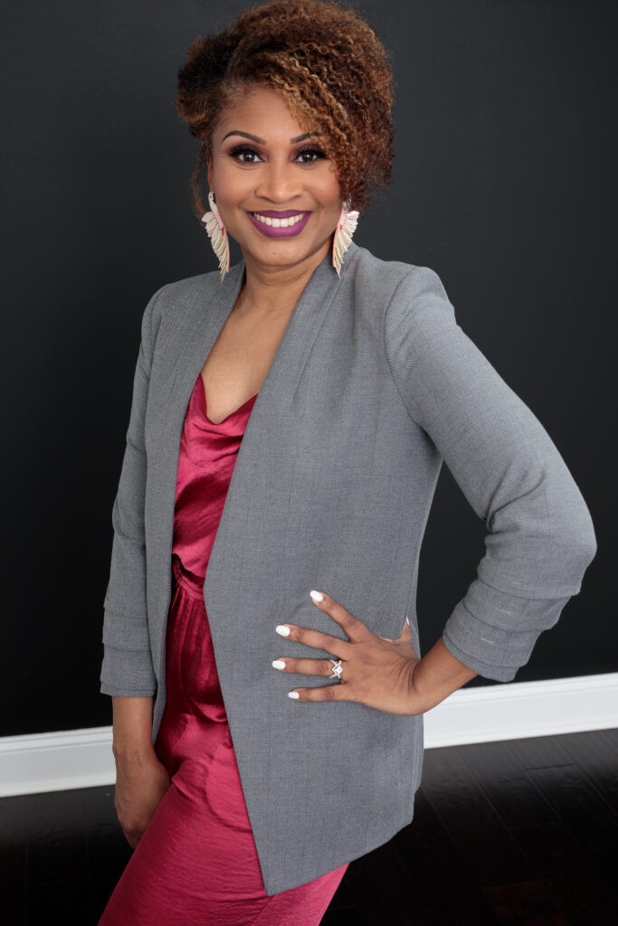 Headshot of Dr. Nika White, an African American woman with short curly brown hair, a fuchsia dress, a grey blazer, and bird-shaped drop earrings, smiling at the camera with one hand on her hip.