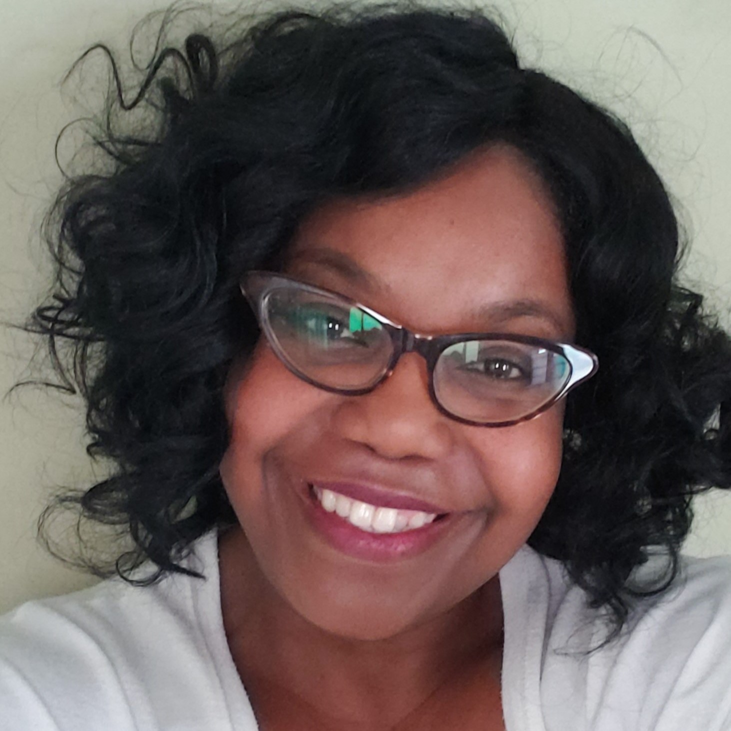 Headshot of Cory, a Black Indigenous woman with short curly black hair, glasses, and a white top smiling at the camera in front of an off-white wall