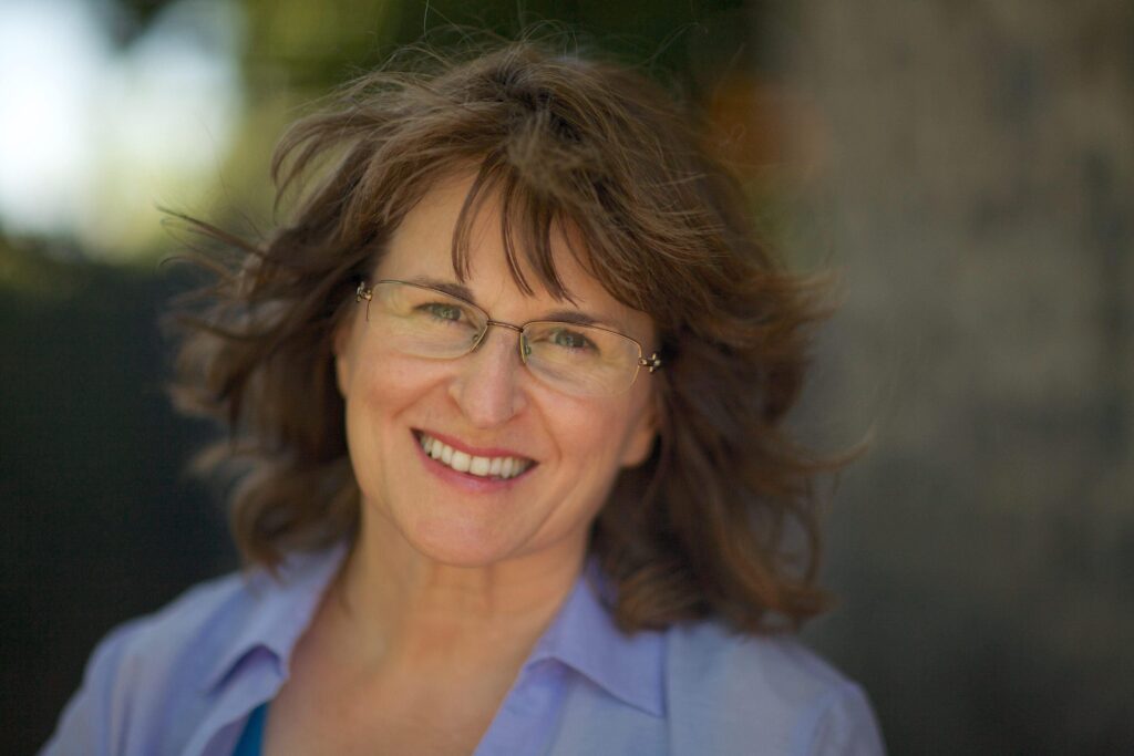 Headshot of Karla McLaren, a smiling, middle-aged, brown-haired, green-eyed, White lady person with glasses and a blue shirt