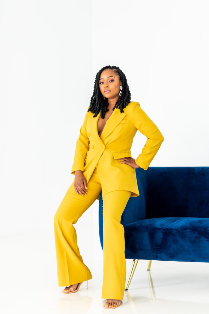 Photo of Sherrell Dorsey, a Black woman with black hair in twists, silver and green statement earrings, and a yellow, standing next to a dark blue couch.