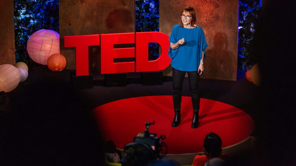 Melinda speaking on the red TED stage in a blue shirt with the large red TED letters in the background