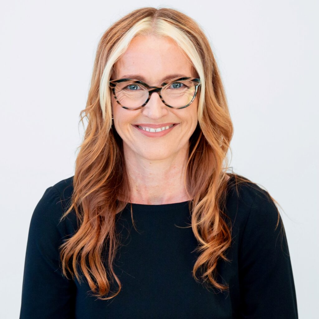 Melinda is smiling in front of a white backdrop looking at the camera. White woman with long red and blonde hair, wearing glasses and a black long sleeved blouse.