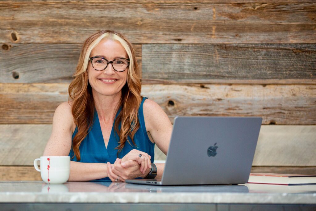 Melinda sits at a table with a coffee cup and Macbook computer. White woman with long red hair, wearing glasses and a sleeveless turquoise shirt. Background is a wood wall.