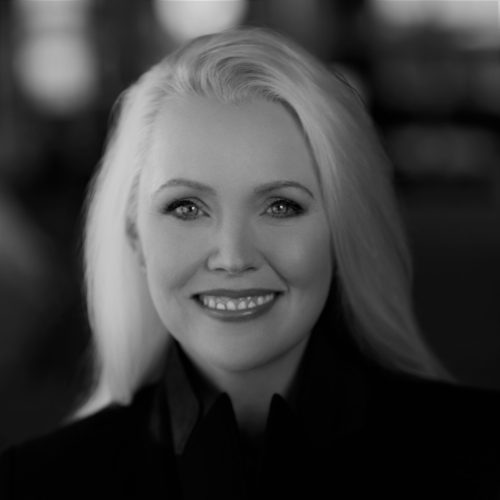 Headshot of Cari Guittard, a White, cis gender female photographed in black and white with long blonde hair and dark button up shirt smiling at the camera with a dark blurry background