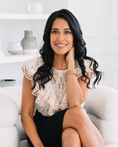 Headshot of Ritu Bhasin, a South Asian woman with long curled black hair, nose ring, white lace blouse, black pencil skirt, and gold wristwatch smiling at the camera and sitting on a white couch with her knees crossed and one elbow on her knee with her hand resting on her neck in front of a white room with light-colored decor.