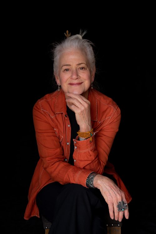 Headshot of Ellen Petry Leanse, a light-skinned female elder with white hair in an updo, black top, black pants, orange jacket, silver dangling earrings, statement bracelets, and a statement ring looking at the camera and sitting on a stool leaning her elbows forward on her legs in front of a black backdrop